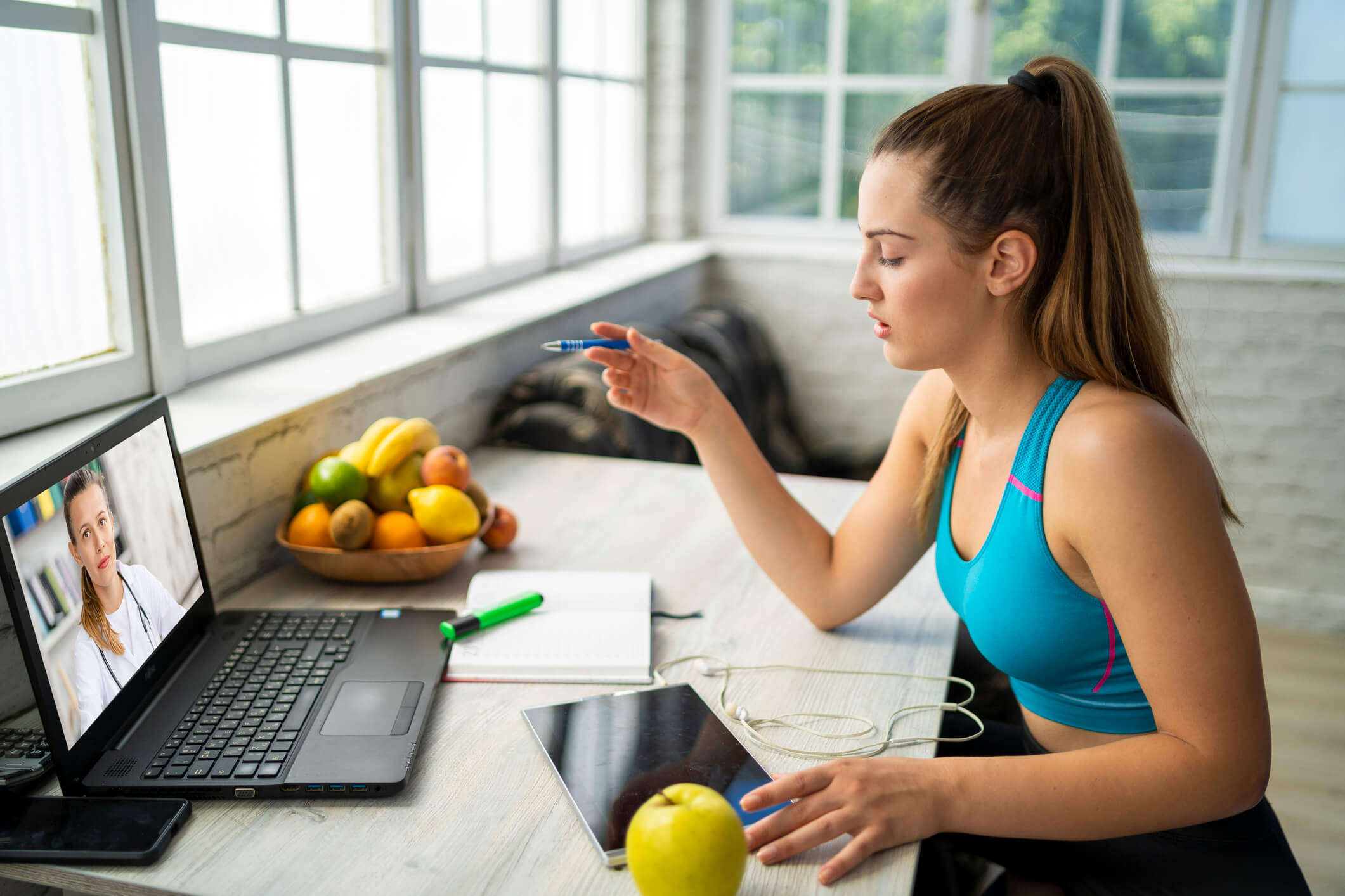 Young woman sitting at a desk with fresh fruit on it talking to a doctor about the FAQs she has regarding her weight loss plan.