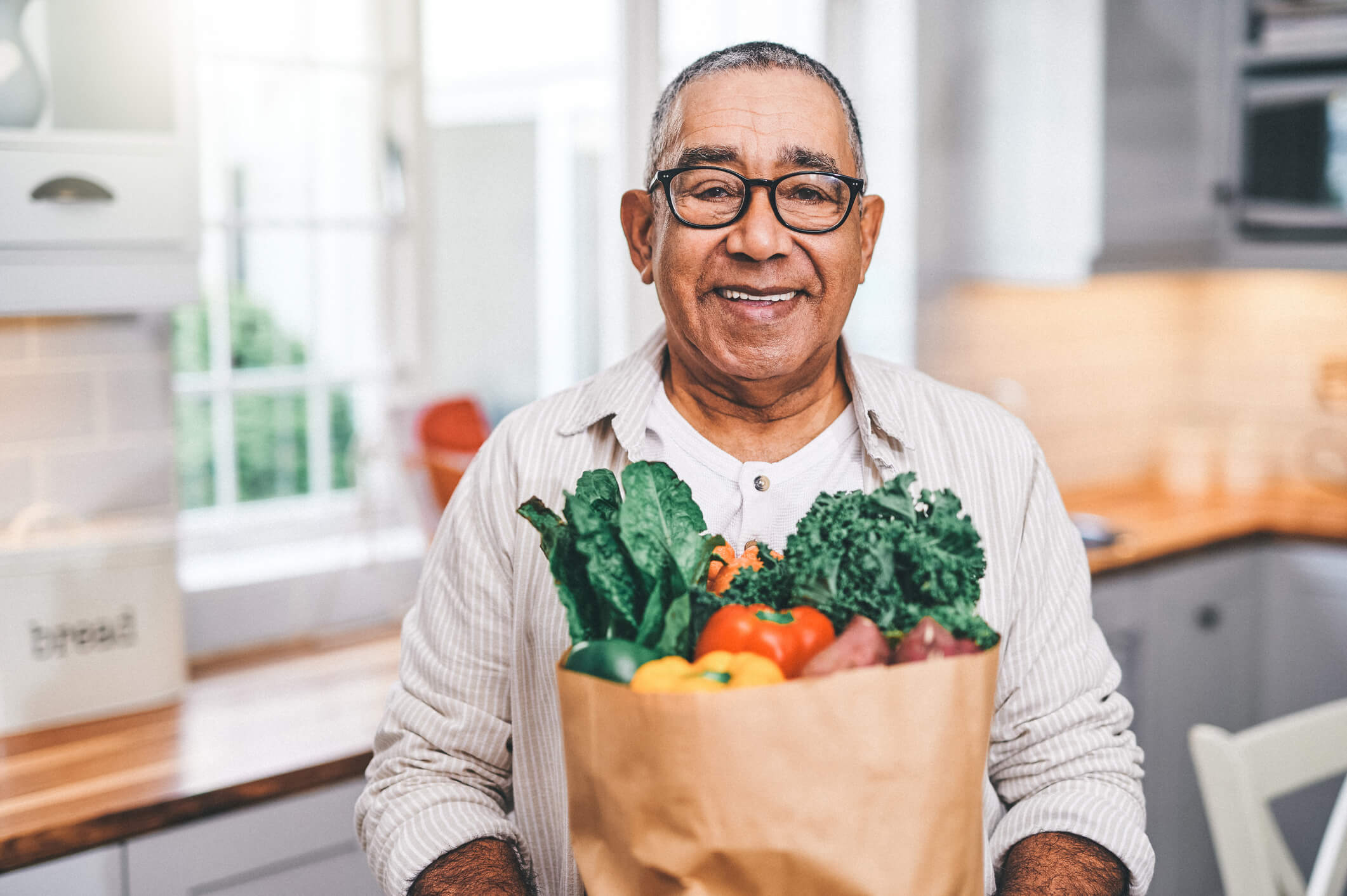 Older man with glasses carrying a brown paper bag full of fresh fruits in vegetables while standing in his modern kitchen.