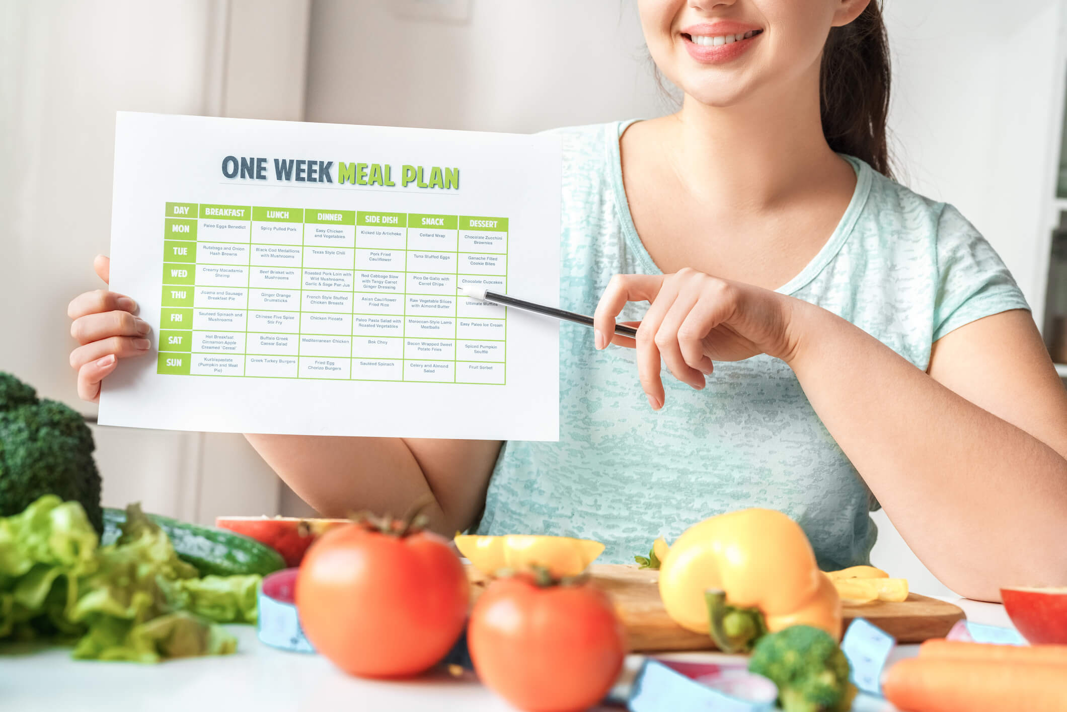 Woman pointing at printed out weight loss nutrition plans with a pen in front of a table full of fresh vegetables.