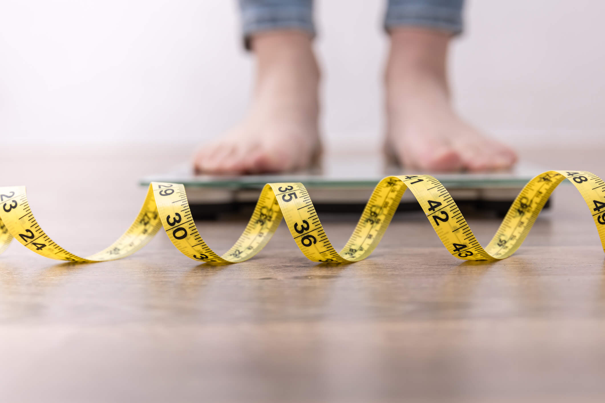 Feet of someone following weight loss programs on a scale behind a coiled measuring tape on the ground.