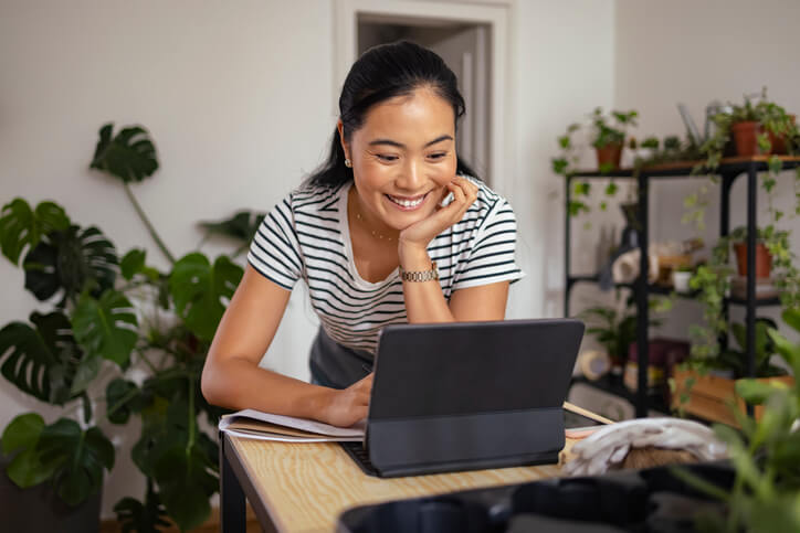 woman in a striped shirt surrounded by plants smiling at her computer screen while talking to the doctor at Advanced Weight Loss Clinic.