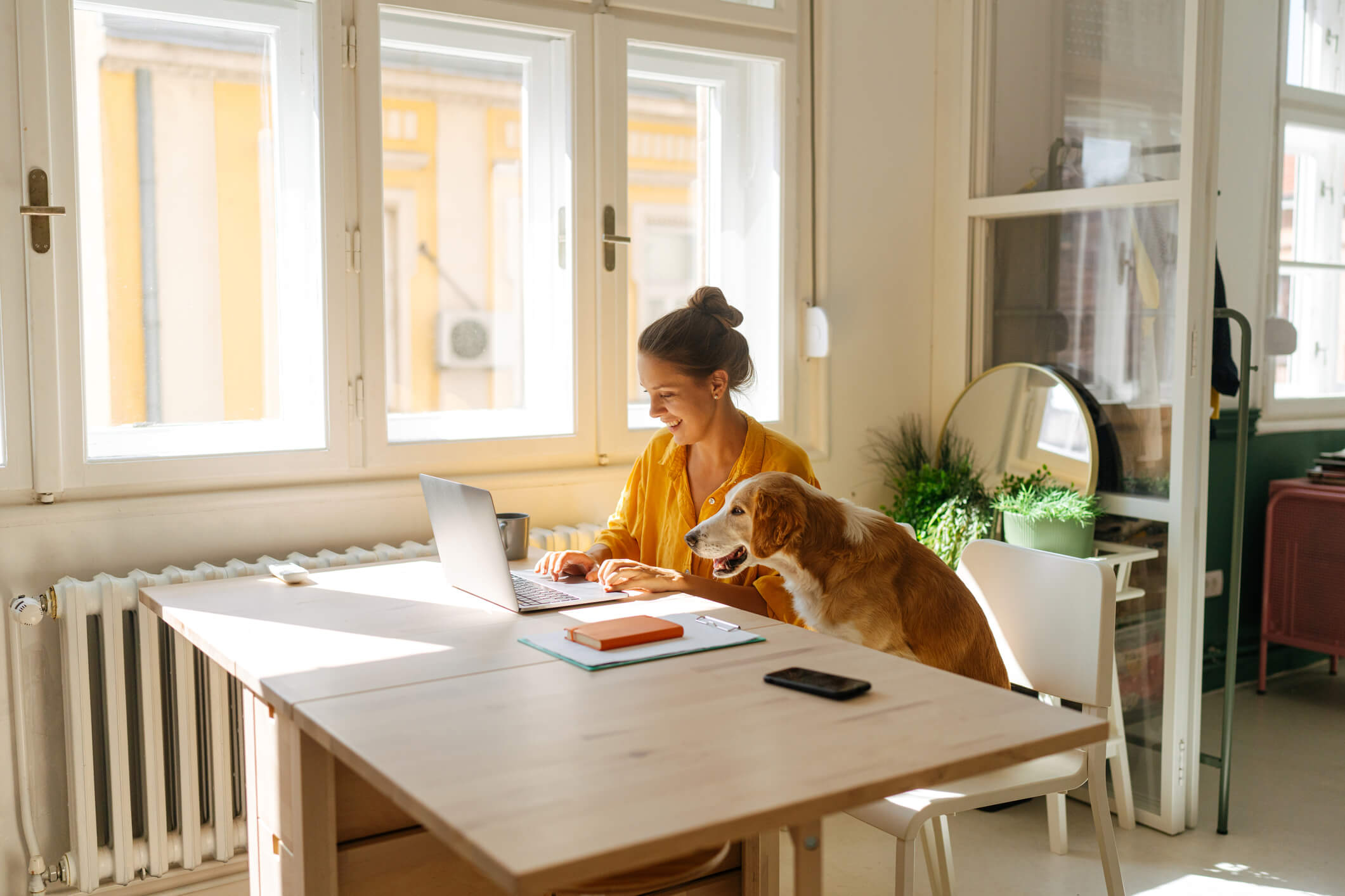 Woman sitting at wooden table with golden retriever next to her looking at her laptop to log into her telehealth appointment.